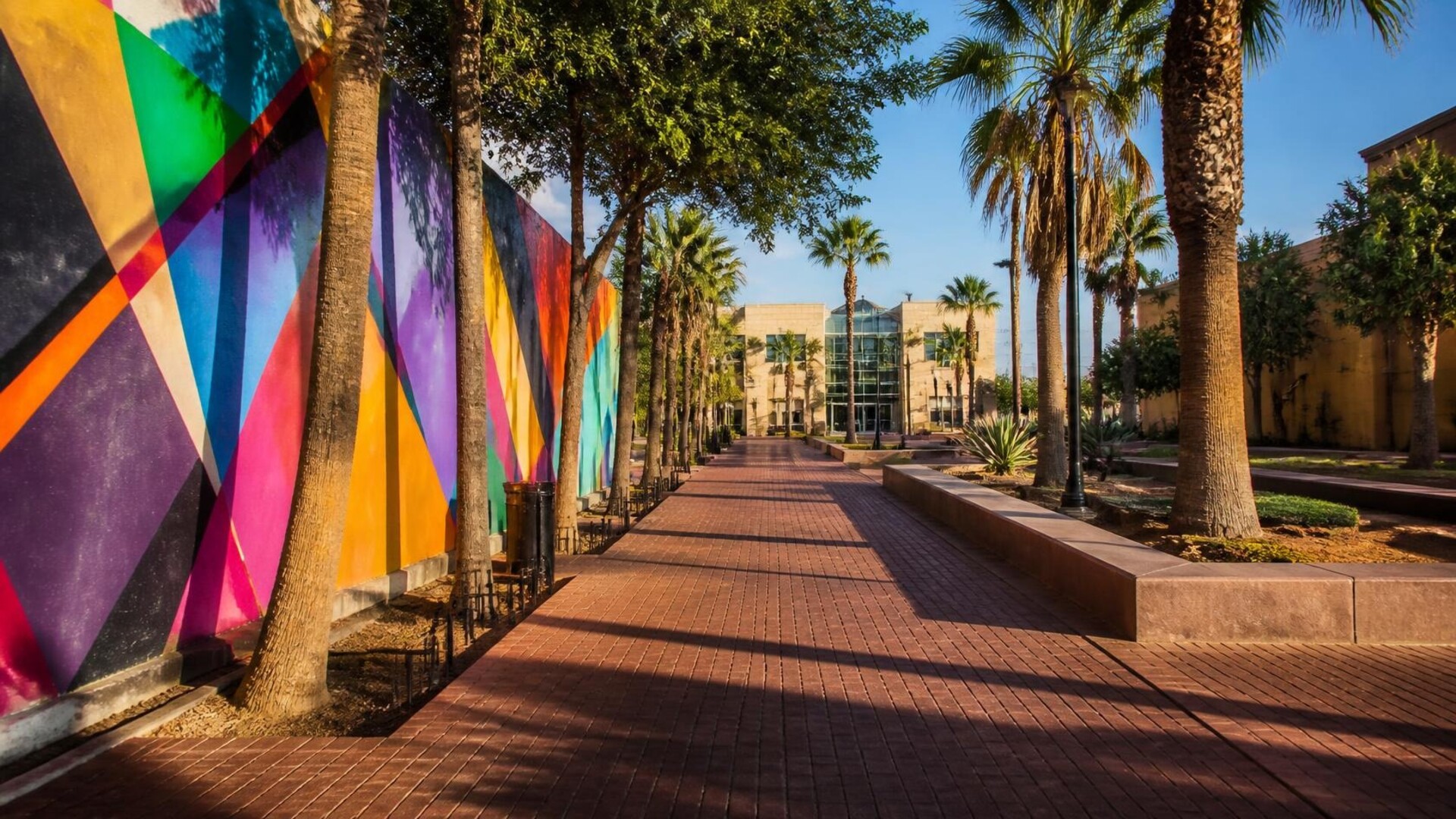Sunny South Texas cultural district street with colorful abstract outdoor mural, palm trees, warm golden light, historic sidewalk and modern cultural center entrance in the distance
